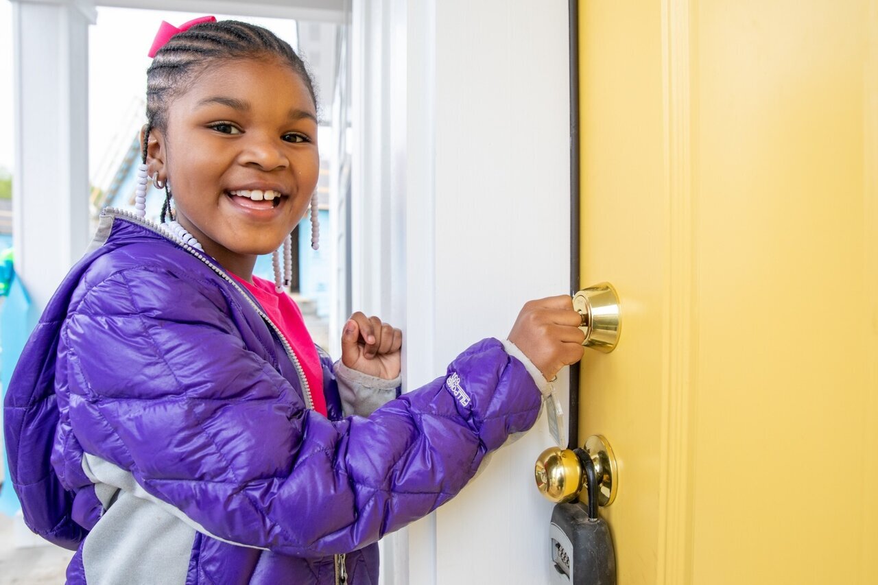 A young girl with braided hair and a bright pink bow, wearing a purple jacket, smiles as she opens a yellow door with a key. She is standing in front of a newly constructed home, symbolizing the joy and security of homeownership.
