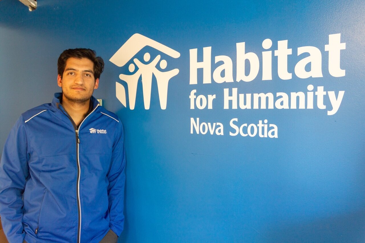 A young man wearing a blue Habitat for Humanity jacket stands smiling in front of a blue wall with the Habitat for Humanity Nova Scotia logo prominently displayed. The logo features the organization’s name and a symbol of a house with three people underneath it.