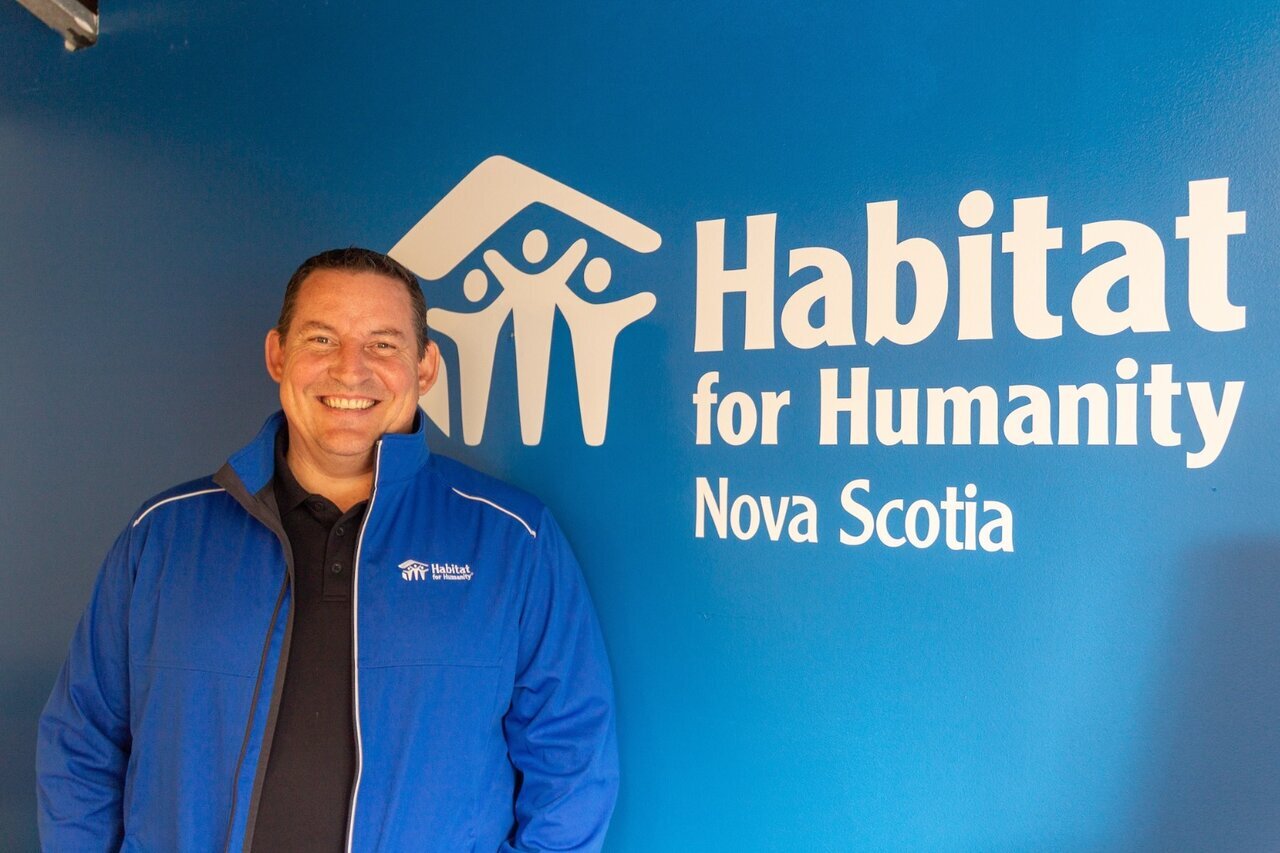 A man in a blue Habitat for Humanity jacket stands smiling in front of a blue wall with the Habitat for Humanity Nova Scotia logo prominently displayed. The logo features the organization’s name and a symbol of a house with three people underneath it.