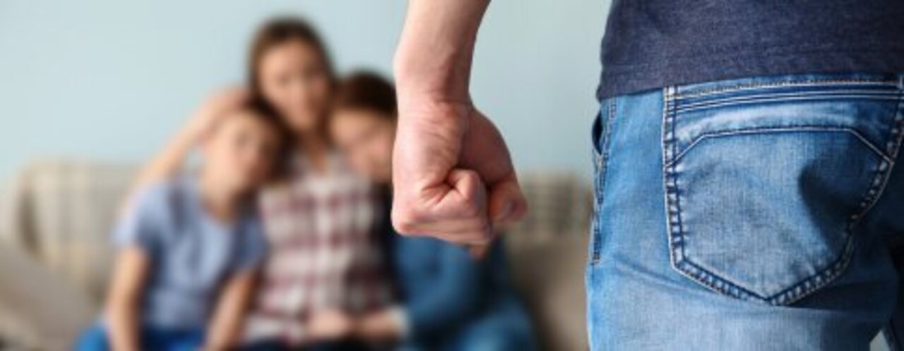 Image shows man standing, fist clenched, facing a blured out women and children who are clutching each other