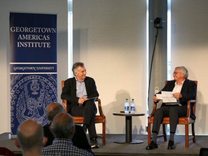 Mauricio Marci and Moises Naim sit in brown chairs on a stage with a blue banner to the left.