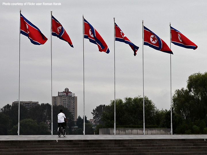 Six tall North Korean flag poles next to each other as man rides his bicycle below them (via Flickr user Roman Harak)