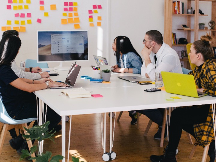 People sitting around a table with laptops and books, looking at flat screen and sticky notes on wall.