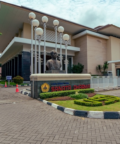 External image of Sanata Dharma University's campus, with a statue, landscaping, and the main building.
