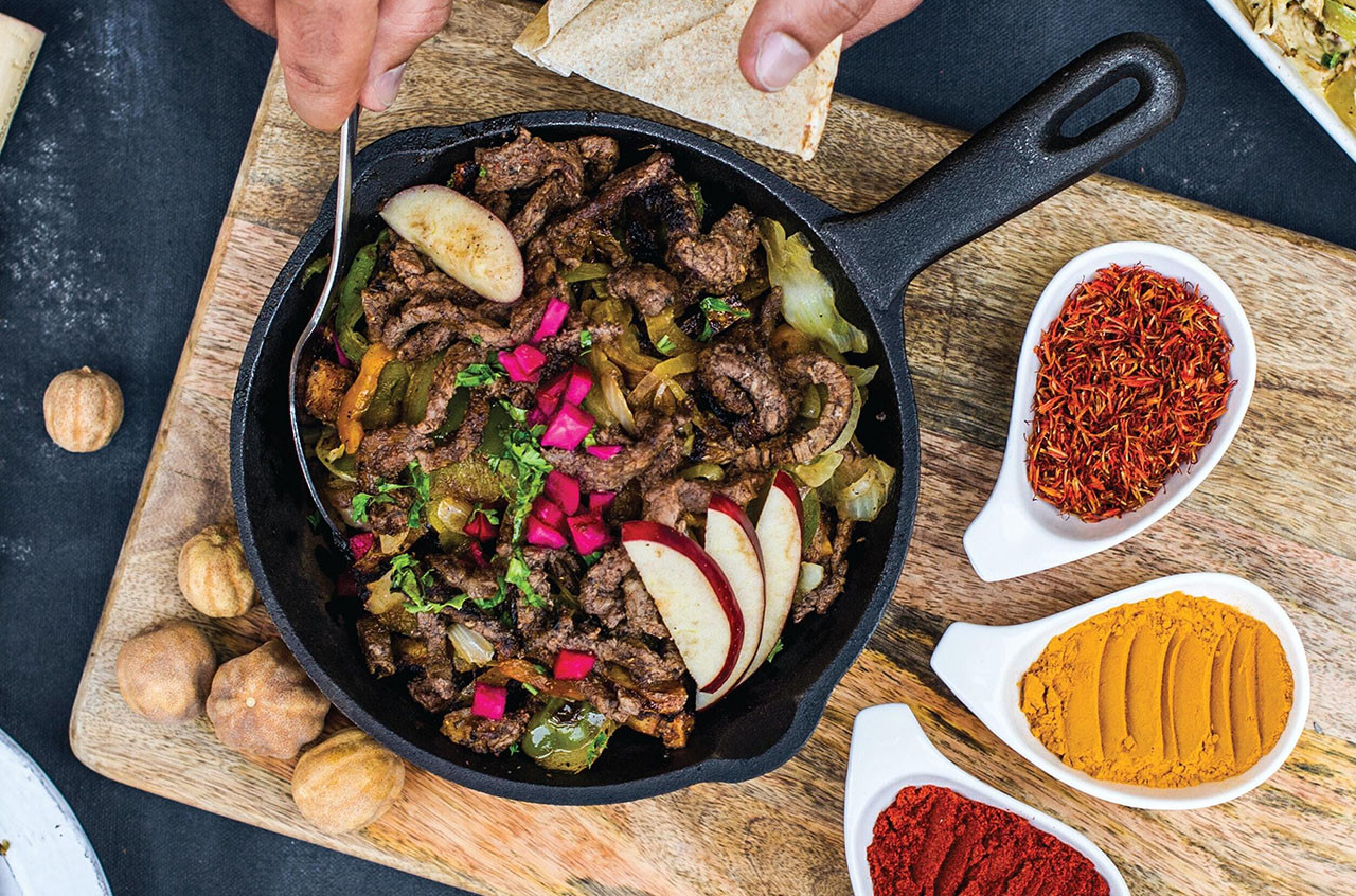 An assortment of meat and vegetables in a cast iron pan, surrounded by white containers of bright spices, on a wooden cutting board.