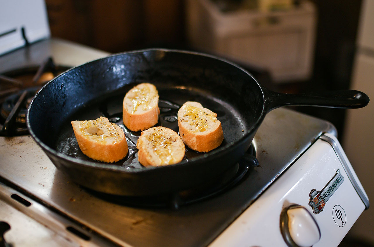 Slices of baguettes with olive oil on them, sitting in a cast iron pan on an oven.