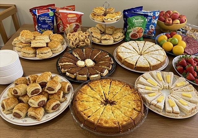 A table full of buffet food like crisps, sausage rolls and sandwiches.
