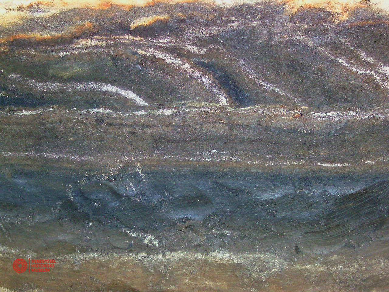 For det utrænede øje kunne dette her være et hvilket som helst oliemaleri, men en arkæolog ser straks, at det er pløjespor! De hvide striber er sand. jernaldergrav