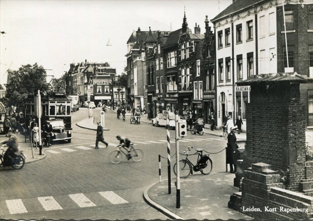 In Leiden kijken we naar verkeersdrukte op het Kort Rapenburg. De A325 van de stadsdienst staat op het punt om linksaf de Breestraat in te rijden. De nieuw geïnstalleerde verkeerslichten laat het verkeer echter nog even wachten. Een nog jonge Joop Zoetemelk wist overigens nog net aan het oranje licht op de Breestraat te ontsnappen. De verkeersagent, met witte platte pet, die voorheen een klapbord bediende om het verkeer te regelen maakt nu gebruik van een geavanceerd schakeltableau, rechts op de foto. Prentbriefkaart van rond 1960, fotograaf onbekend