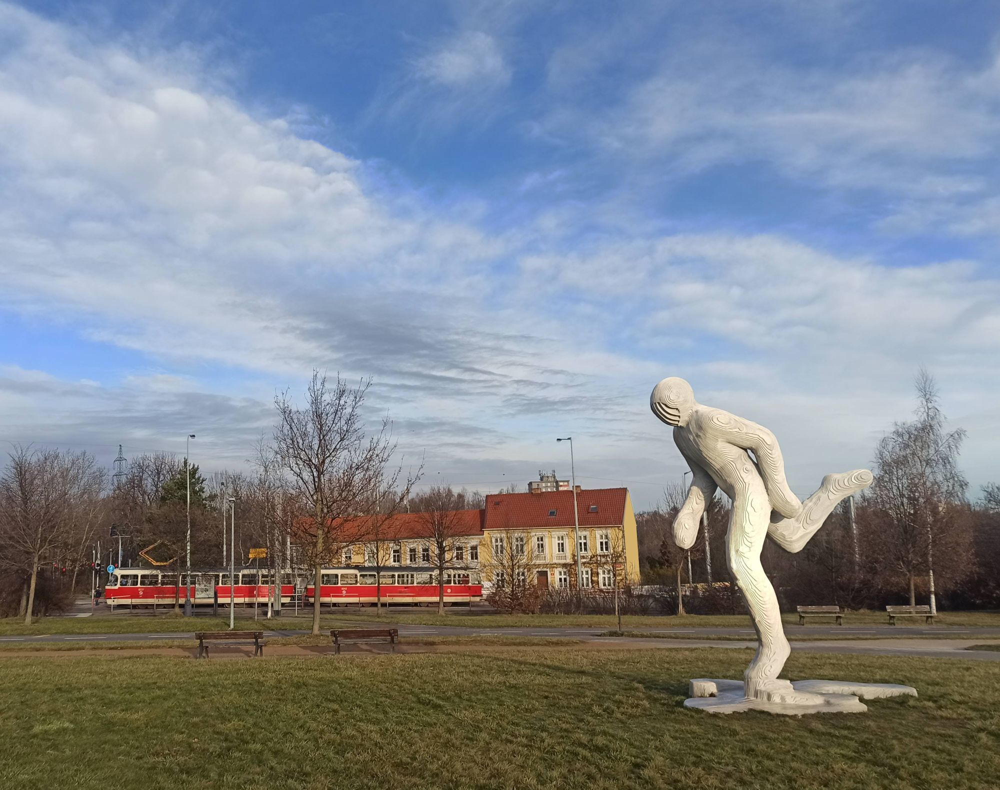 Foto sochy Ocelový Běžec za slunečného zimního rána s tramvají T3 v pozadí.