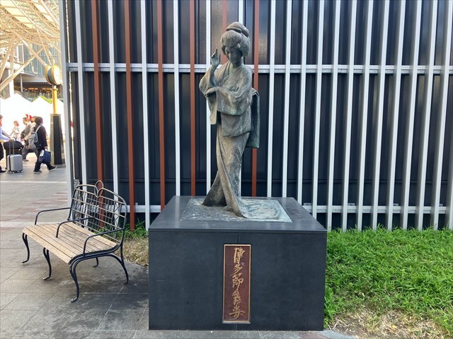 博多人形の銅像の写真　人形は着物姿の女性で右手を右頬の横に、左手は右肘につけている