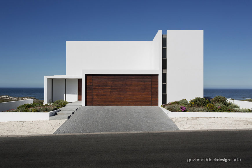 As seen from the street, the home conceals most of its details behind a broad swath of white, with only a vertical window strip and dark wood garage door breaking up the facade. As seen from the street, the home conceals most of its details behind a broad swath of white, with only a vertical window strip and dark wood garage door breaking up the facade.