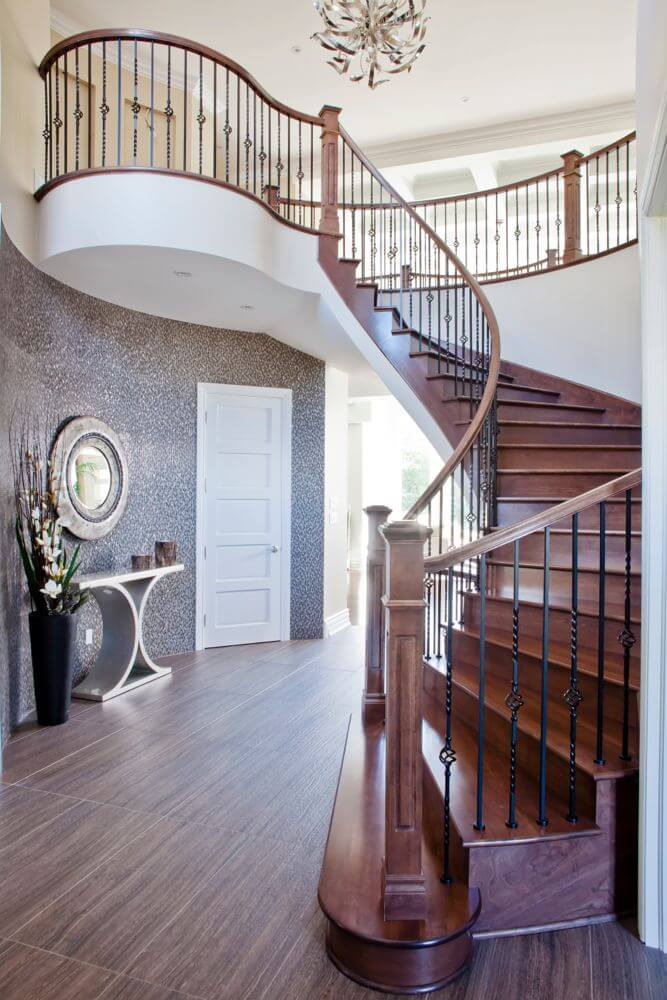 Main staircase is a beautifully curved dark wood design, with wrought-iron railing supports. Landing at a dark hardwood floor, this central hall space features artful metallic decorative table and mirror against patterned wall, while daylight beams in through large living room windows in background. Main staircase is a beautifully curved dark wood design, with wrought-iron railing supports. Landing at a dark hardwood floor, this central hall space features artful metallic decorative table and mirror against patterned wall, while daylight beams in through large living room windows in background.