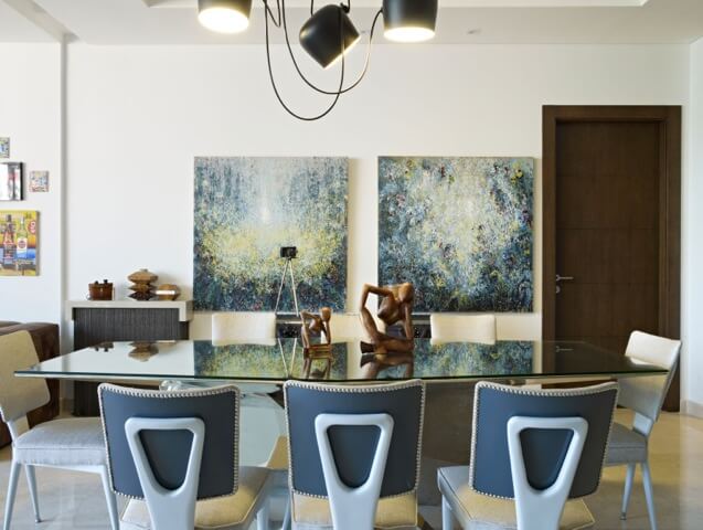 Dining room set, with immense glass-top table standing beneath black recycled component chandelier, holds the pair of aforementioned paintings next to dark natural wood door. Dining room set, with immense glass-top table standing beneath black recycled component chandelier, holds the pair of aforementioned paintings next to dark natural wood door.