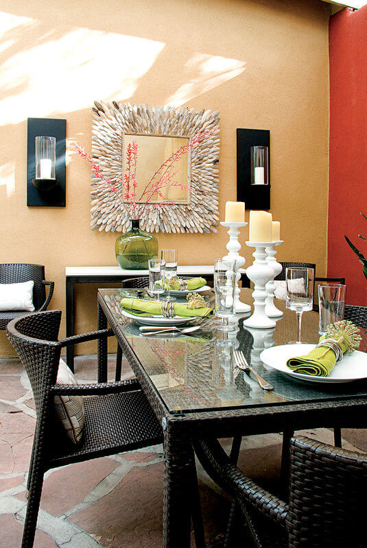 This patio dining space mimics a traditional indoor space, replete with sconces on the stucco wall, flanking a large decorative mirror. Resin wicker seating and glass topped table complete the dining set. This patio dining space mimics a traditional indoor space, replete with sconces on the stucco wall, flanking a large decorative mirror. Resin wicker seating and glass topped table complete the dining set.