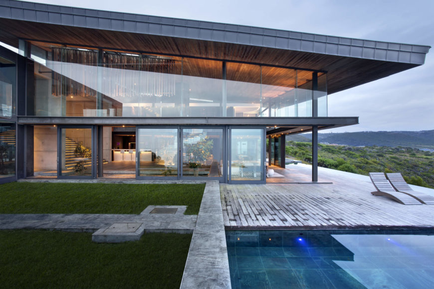 The patio wraps the home with wood planks, a stone path, and small lawn next to the pool. We see the sloped roof hovering above the glass wrapped structure, offering unobscured views all around. The patio wraps the home with wood planks, a stone path, and small lawn next to the pool. We see the sloped roof hovering above the glass wrapped structure, offering unobscured views all around.