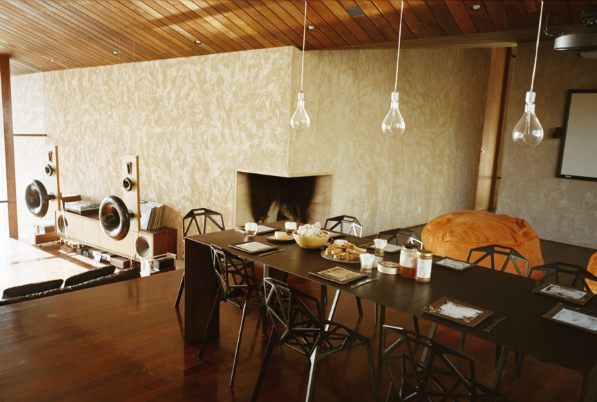 The lengthy contemporary dark wood dining table is complemented by a set of geometric, minimalist dining chairs below a trio of bare bulb pendant lights. A corner fireplace is carved into the wall, while the elaborate music center can be seen at left. The lengthy contemporary dark wood dining table is complemented by a set of geometric, minimalist dining chairs below a trio of bare bulb pendant lights. A corner fireplace is carved into the wall, while the elaborate music center can be seen at left.