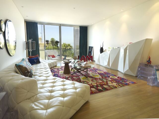 The spacious living room has many components. The focal point of the room is the largest piece of furniture: the couch. The couch is a custom tufted sectional in white that dominates the left half of the room. On the right wall is a unique geometric shelf with concave and convex angles. A colorful patterned rug adds a punch of color to the mostly white and neutral room. The spacious living room has many components. The focal point of the room is the largest piece of furniture: the couch. The couch is a custom tufted sectional in white that dominates the left half of the room. On the right wall is a unique geometric shelf with concave and convex angles. A colorful patterned rug adds a punch of color to the mostly white and neutral room.