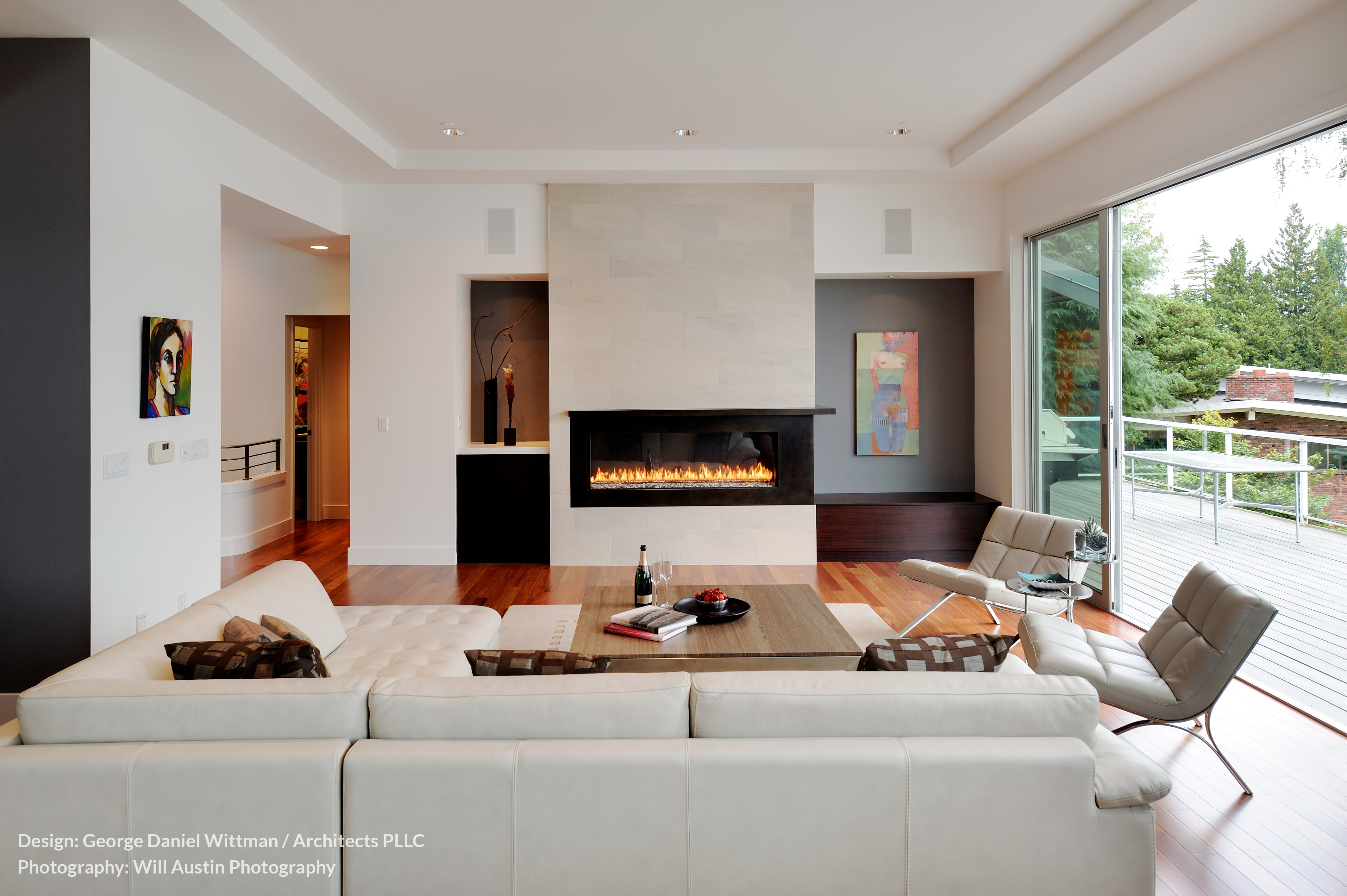 The living room opens almost entirely to the upper balcony, with large sliding glass panels as seen at right. A modern gas fireplace dominates the view, while white leather gracing the sectional and chairs contrasts with rich hardwood flooring. The living room opens almost entirely to the upper balcony, with large sliding glass panels as seen at right. A modern gas fireplace dominates the view, while white leather gracing the sectional and chairs contrasts with rich hardwood flooring.