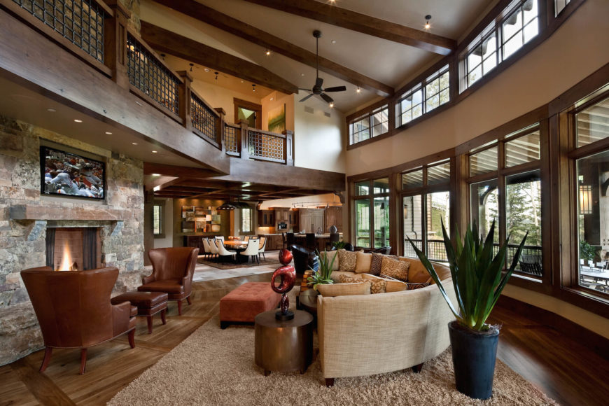 In the sprawling, open design living room, we see a full two stories of windows reaching toward the exposed wood beam ceiling, allowing natural light to spread across the expanse of hardwood flooring. A large rounded sectional stands at center before the stone fireplace, with catwalk hallway overhead. In the sprawling, open design living room, we see a full two stories of windows reaching toward the exposed wood beam ceiling, allowing natural light to spread across the expanse of hardwood flooring. A large rounded sectional stands at center before the stone fireplace, with catwalk hallway overhead.