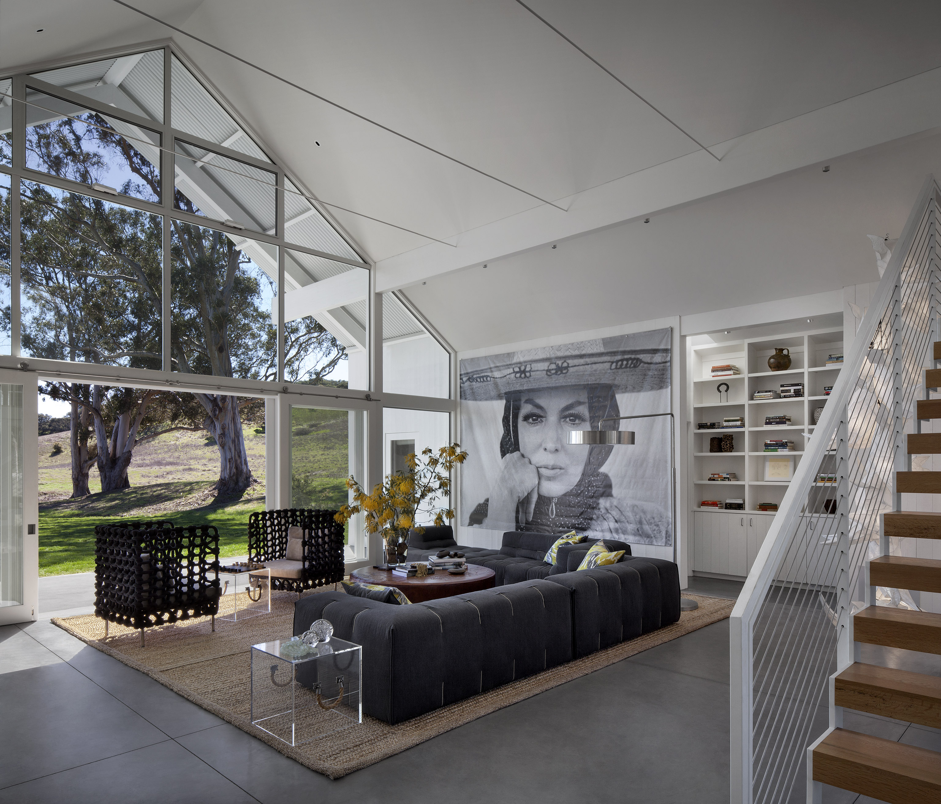 The living room is flush with unique touches, from the large button tufted sectional and a pair of knotted-frame club chairs to the immense black and white portrait standing next to built-in wall shelving. A large woven area rug centers the furniture over the concrete flooring. The living room is flush with unique touches, from the large button tufted sectional and a pair of knotted-frame club chairs to the immense black and white portrait standing next to built-in wall shelving. A large woven area rug centers the furniture over the concrete flooring.