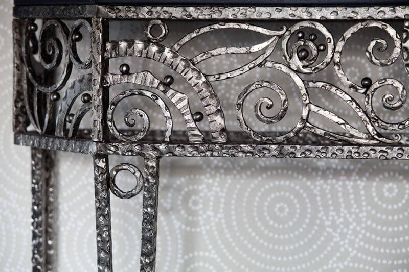 The small entryway into the apartment has an ornate wrought iron side table against a light gray wallpaper with white concentric circles. The small entryway into the apartment has an ornate wrought iron side table against a light gray wallpaper with white concentric circles.