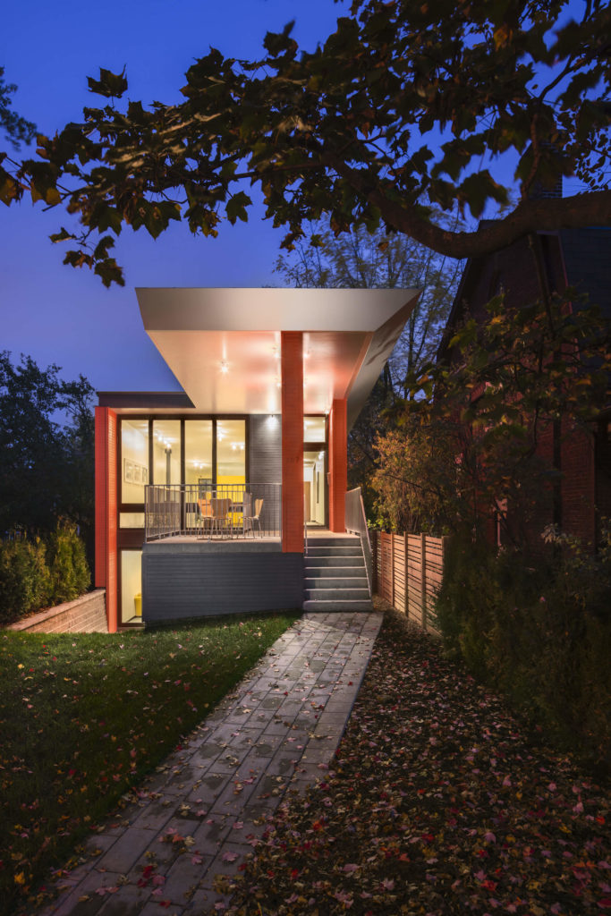 Viewed at nighttime, the interior lighting glows outward via the full height glass panels covering the front of the home. A large front patio stands sheltered beneath the extended rooftop. Viewed at nighttime, the interior lighting glows outward via the full height glass panels covering the front of the home. A large front patio stands sheltered beneath the extended rooftop.