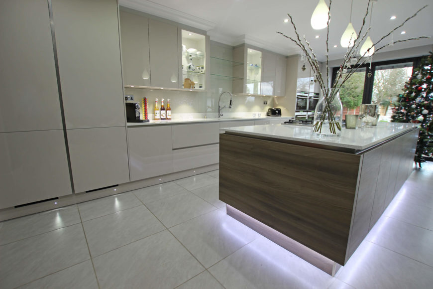 The island visually floats over the large format tile flooring, with a series of lighting strips. The dark wood grain adds contrast to a mostly white space. The island visually floats over the large format tile flooring, with a series of lighting strips. The dark wood grain adds contrast to a mostly white space.