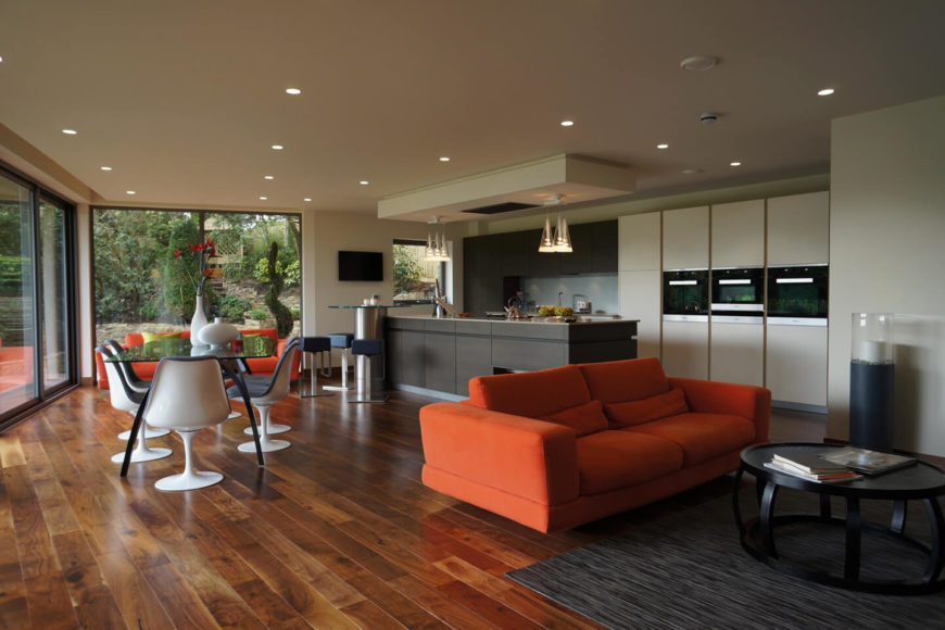 Moving indoors, we see the open-plan space at the heart of the home, containing kitchen, dining, and a living room area over rich hardwood flooring. The sleekly modern kitchen sports dark toned wood and white cabinetry, while the black glass dining table and bright orange living room sofa add bold contrast and color. Moving indoors, we see the open-plan space at the heart of the home, containing kitchen, dining, and a living room area over rich hardwood flooring. The sleekly modern kitchen sports dark toned wood and white cabinetry, while the black glass dining table and bright orange living room sofa add bold contrast and color.