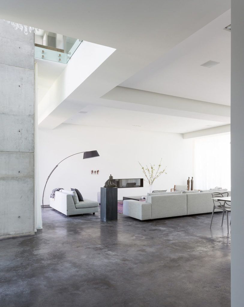 The grand, open-plan living room features a set of light grey contemporary sectionals over an expanse of dark stone flooring, contrasting with bright white surroundings. The upper floor can be seen peeking over at top left. The grand, open-plan living room features a set of light grey contemporary sectionals over an expanse of dark stone flooring, contrasting with bright white surroundings. The upper floor can be seen peeking over at top left.