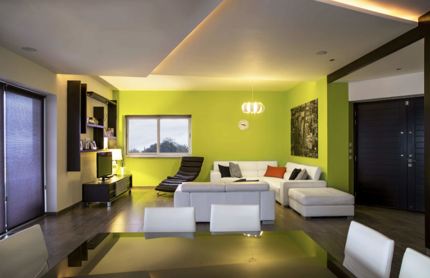 Viewed from across the deep black dining table, the living room stands hugged by a bold lime green set of walls, distinguishing a playful space from the rest of the home interior. Viewed from across the deep black dining table, the living room stands hugged by a bold lime green set of walls, distinguishing a playful space from the rest of the home interior.