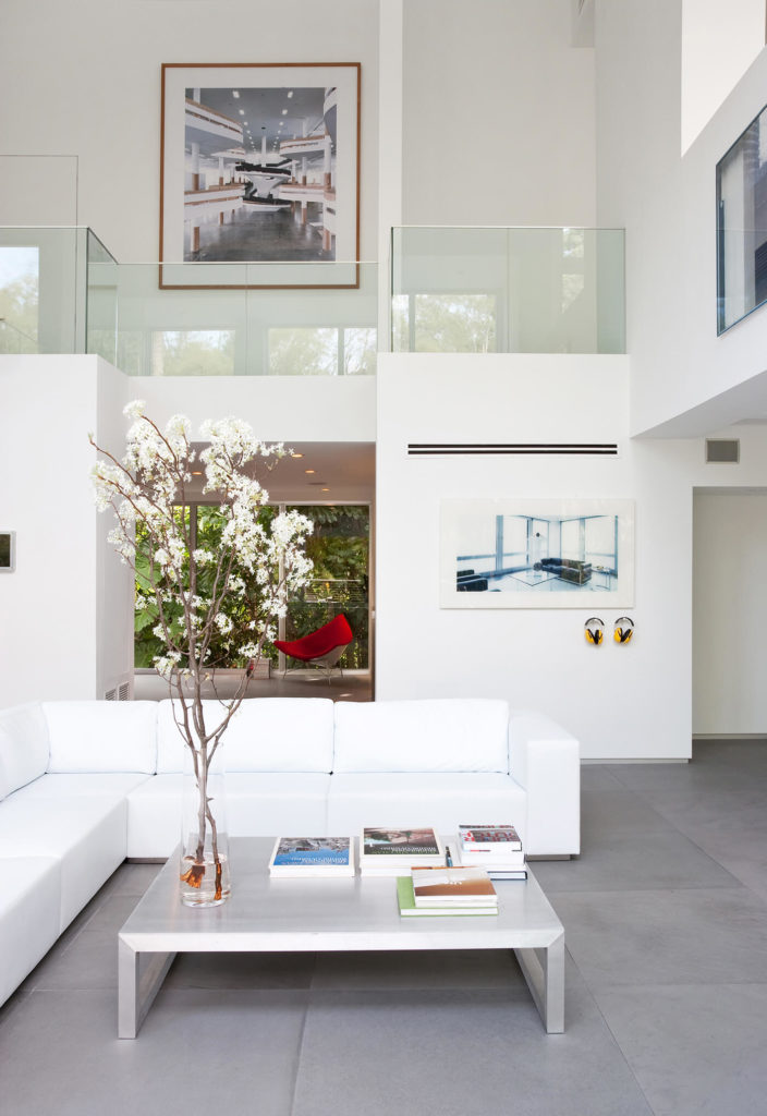 The open-plan, two-story living room centers on a large L-shaped sectional in bright white, hugging an immense metallic coffee table. Loft-style second floor space overlooks the room, with seamless glass walls for safety and clear visual space. The open-plan, two-story living room centers on a large L-shaped sectional in bright white, hugging an immense metallic coffee table. Loft-style second floor space overlooks the room, with seamless glass walls for safety and clear visual space.
