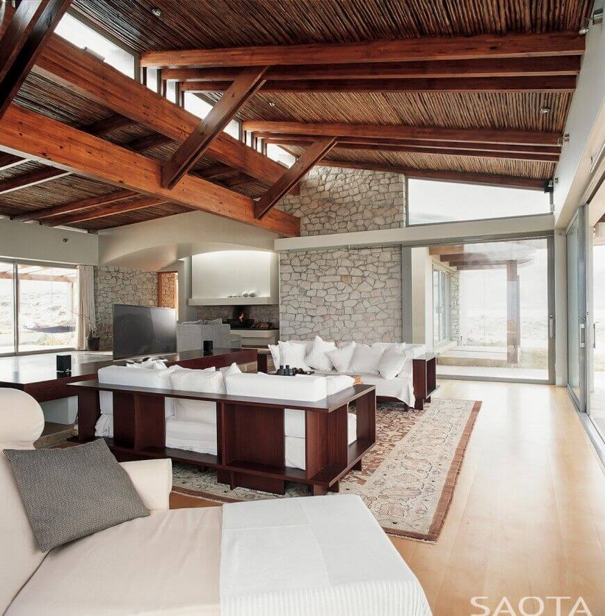An enormously tall stone accent wall soars above the wooden ceiling of this contemporary living room in light neutrals.