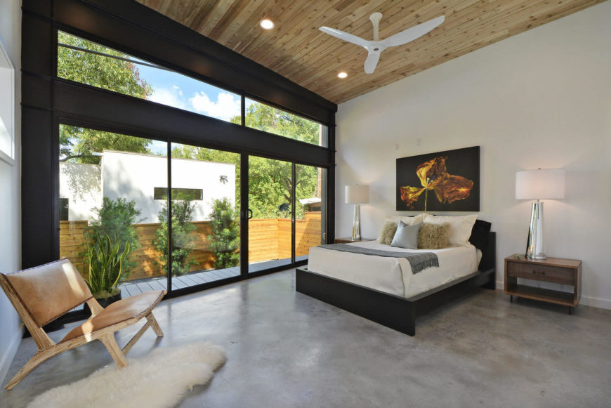 Master bedroom features a vaulted wooden shiplap ceiling with a mounted white fan. It has a light wooden chair accented with a faux fur rug over concrete flooring.