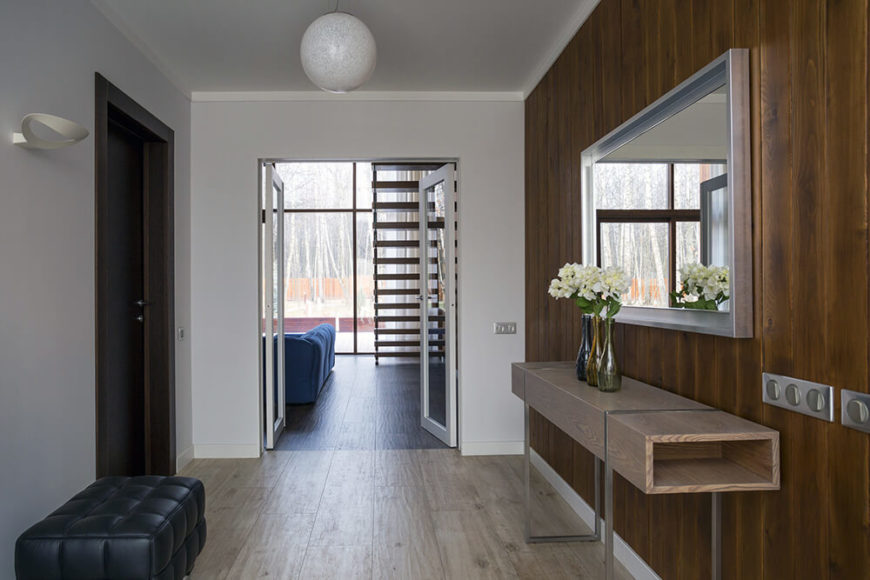 Entry to the home is provided through a wide hall, which features a larch plank accent wall with a minimalist table and a silver-framed mirror. The left wall is starkly minimalist in light gray. Glass double doors lead into the main floor living room. Entry to the home is provided through a wide hall, which features a larch plank accent wall with a minimalist table and a silver-framed mirror. The left wall is starkly minimalist in light gray. Glass double doors lead into the main floor living room.