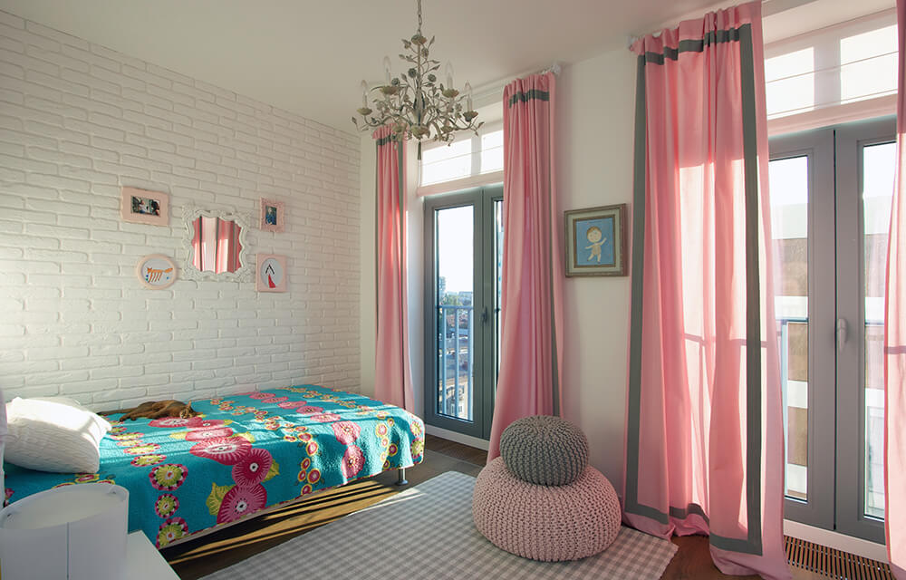 Charming master bedroom with a blue bed placed on the white brick walls. It includes two woven bean bags and floor to ceiling gray paneled windows covered with pink curtains.