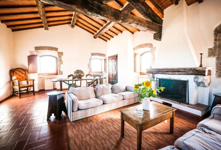 The warm red tile flooring and white walls make for a rich contrast in this living room, beneath a sleek wood panel ceiling with exposed rustic-style beams. The vast room is surrounded by windows and features a large fireplace.