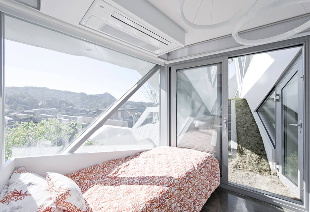 Natural light streams in through the glass windows in this master bedroom. It has a small bed in floral print bedding over a hardwood flooring.
