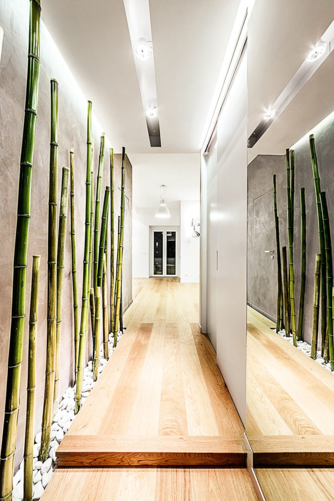 The entry hall introduces the hardwood flooring that informs the entire apartment, flanked by pristine white walls decorated with a slim bamboo forest and a massive mirror. The intricate recessed lighting highlights the length of the bamboo, nested into white stones at the floor. The entry hall introduces the hardwood flooring that informs the entire apartment, flanked by pristine white walls decorated with a slim bamboo forest and a massive mirror. The intricate recessed lighting highlights the length of the bamboo, nested into white stones at the floor.