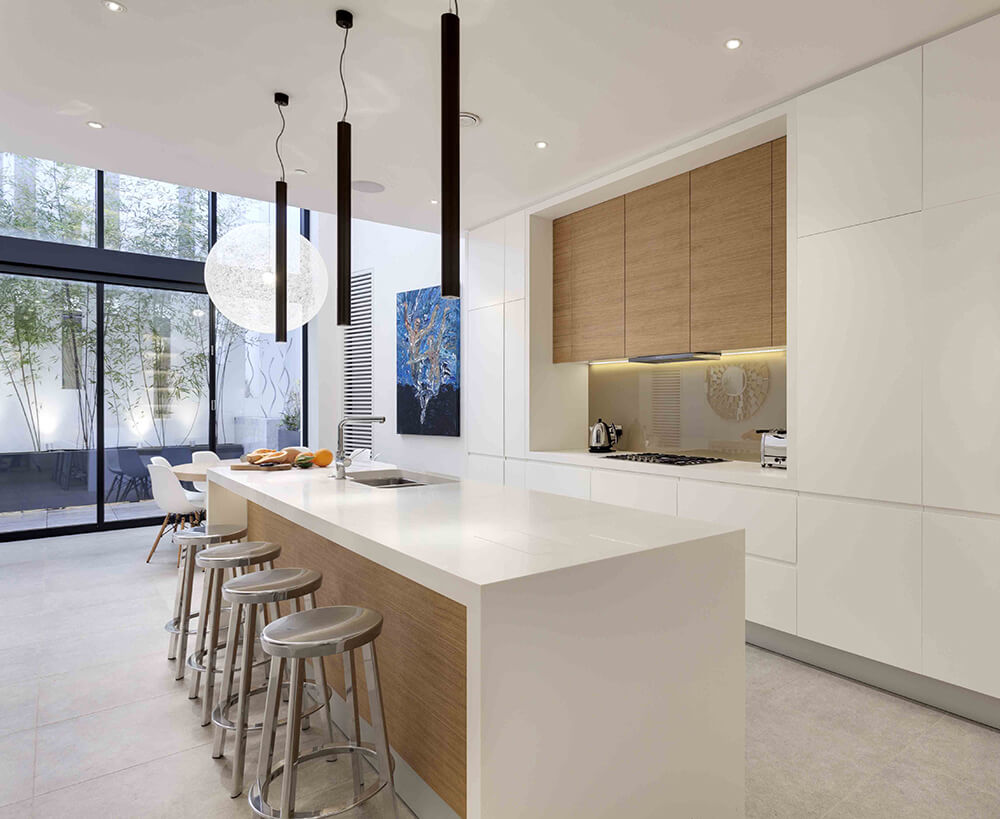 White dine-in kitchen with a breakfast island fitted with a sink and lined with chrome bar stools. It has a wooden vent hood fixed to the glossy backsplash tiles in between sleek cabinetry.