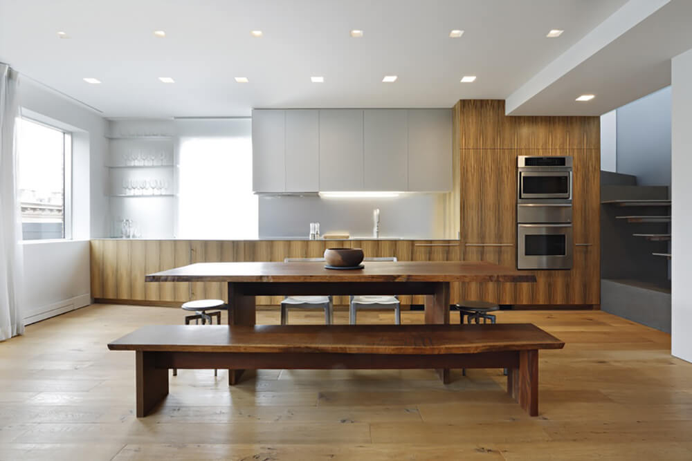 Cozy eat-in kitchen showcases wide wood plank flooring and white ceiling fitted with recessed lights. It has a wooden dining table surrounded with metal chairs and stools along with a wooden bench.