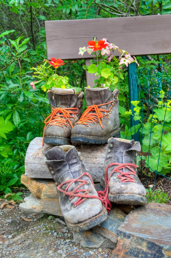 A pair of washed out work boots serve as flower planters while another pair of an even more washed out boots awaits its turn to be revived. The brightly colored laces of both boots add striking contrast to its faded environment.