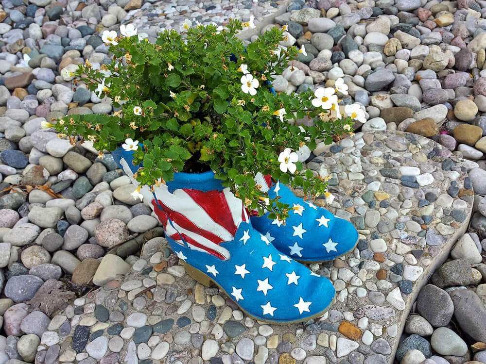 This pair of boots brightly painted with an American flag, standing as planters to white-colored flowers, is a sight to behold in a sea of pebbles and stones.