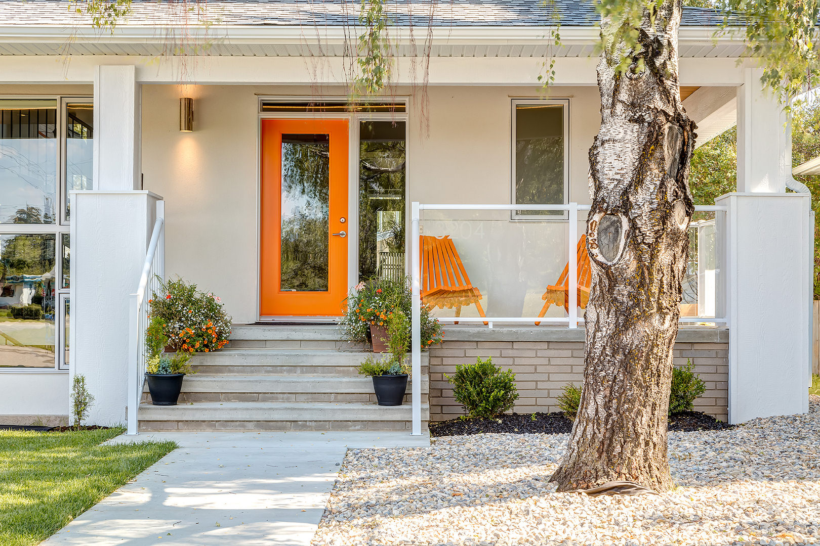 A front yard with concrete pathway ornamented with white pebbles and grass on each side, potted flowers and greenery on the steps. Orange coating on the glass front door and a pair of orange wooded chairs will approach you as you reach the front porch. A front yard with concrete pathway ornamented with white pebbles and grass on each side, potted flowers and greenery on the steps. Orange coating on the glass front door and a pair of orange wooded chairs will approach you as you reach the front porch.