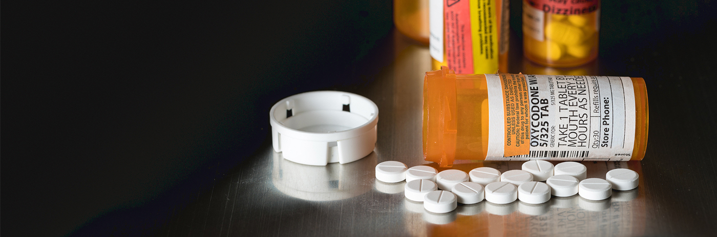 three opioids pill bottles, one of which is laying on its side, sit on a table next to a pile of white pills