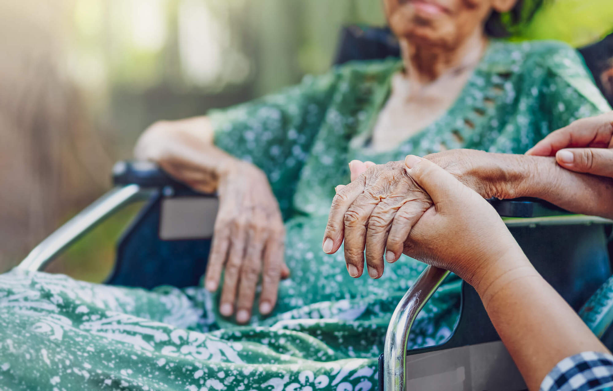 elderly woman wearing a green dress sits in a wheelchair holding hands with an unpictured person