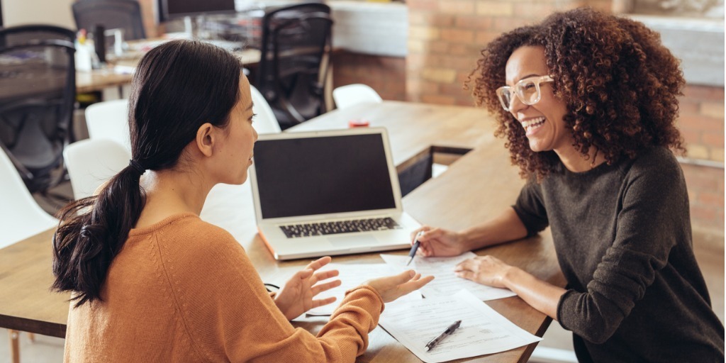 two female colleagues in an exposed brick office space meet at a desk with a laptop and papers
