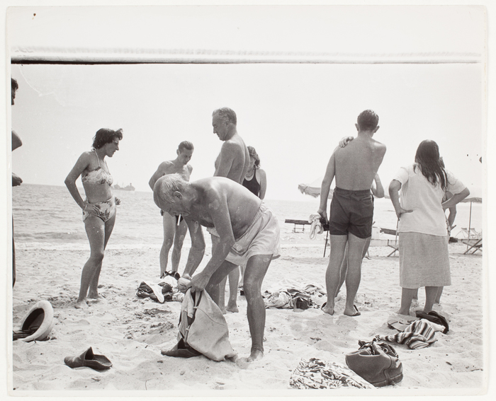 [Pablo Picasso bending, with a cigarette hanging in his lips, and people on the shore, Vallauris, France]