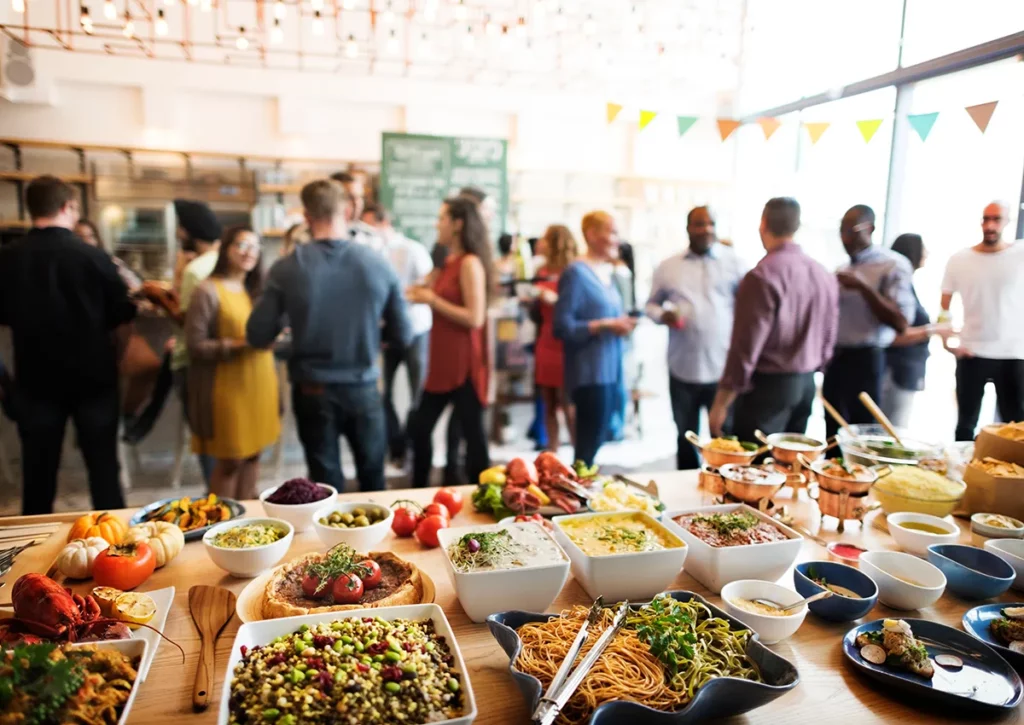 A close-up of a buffet table covered in catered side dishes, snacks, and dips with guests mingling in the background.