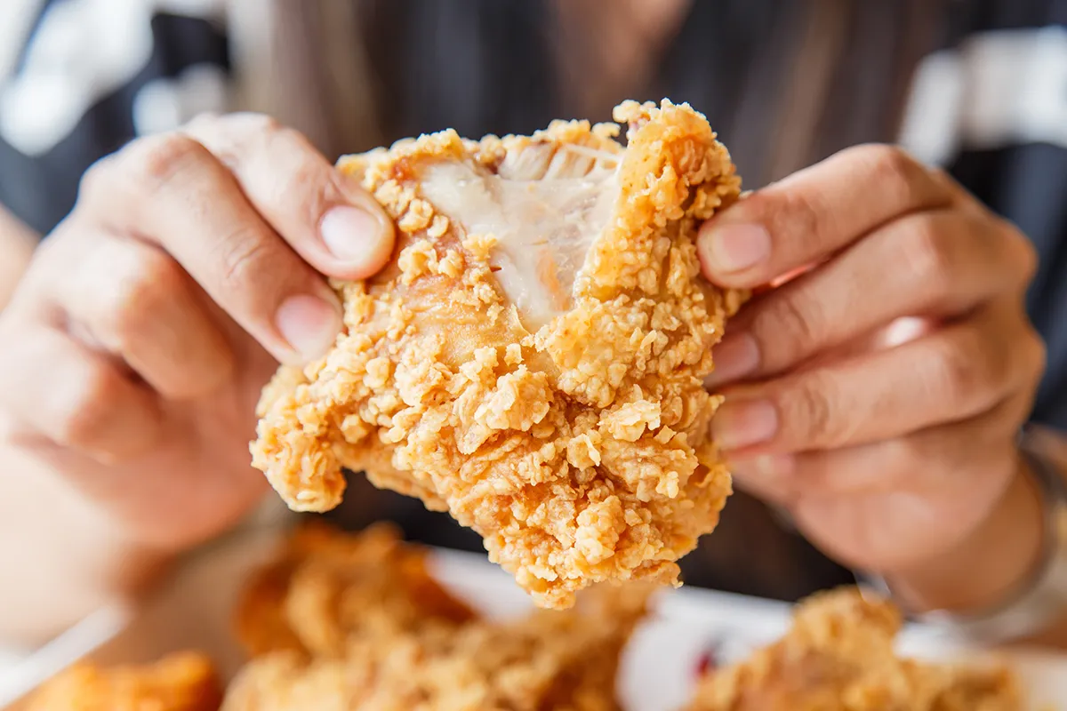 Close-up of hands holding a piece of golden fried chicken with a bite taken out, showing the juicy interior.